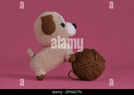 Poupée Amigurumi chiot sur fond rose jouant avec la balle de fil. Un jouet de bricolage doux en coton naturel et laine. Petit chien brun crocheté, handm Banque D'Images