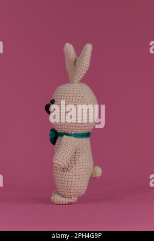 Symbole de Pâques catholique. Lapin gris mignon de Pâques avec noeud papillon vert, crocheté, art fait à la main Vue latérale. Poupée de lapin Amigurumi sur fond rose. SOF Banque D'Images