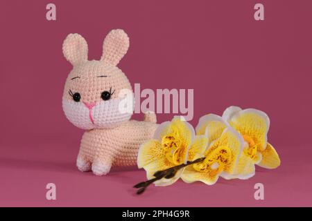 Poupée de lapin Amigurumi sur fond rose avec fleurs d'orchidées jaunes. Jouet bricolage doux en coton et laine naturelle. Symbole du concept de Pâques catholique. Banque D'Images