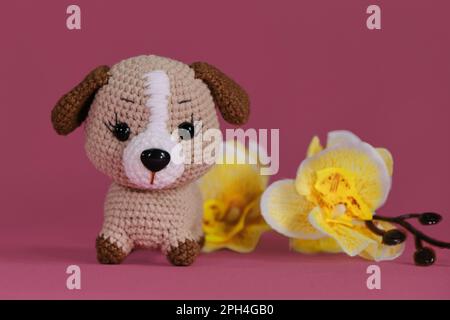 Poupée de chiot Amigurumi sur fond rose avec fleurs d'orchidées jaunes. Jouet bricolage doux en coton naturel et laine. Petit chien brun crocheté, fait à la main Banque D'Images