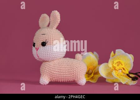 Poupée de lapin Amigurumi sur fond rose avec fleurs d'orchidées jaunes. Jouet bricolage doux en coton et laine naturelle. Symbole du concept de Pâques catholique. Banque D'Images