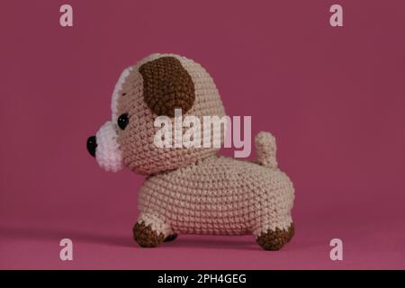 Petit chien brun crocheté, art fait à la main Poupée Amigurumi chiot sur fond rose. Un jouet de bricolage doux en coton naturel et laine. Vue latérale. Banque D'Images