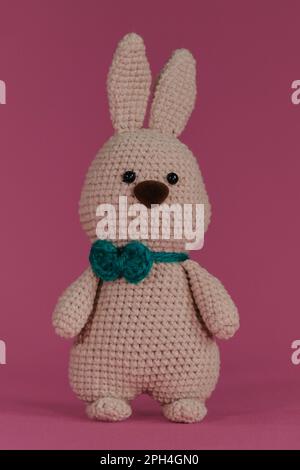 Symbole de Pâques catholique. Lapin gris mignon de Pâques avec noeud papillon vert, crocheté, art fait à la main Poupée de lapin Amigurumi sur fond rose. Jouet bricolage doux m Banque D'Images