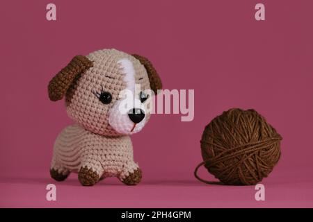 Poupée de chiot Amigurumi sur fond rose à côté de la boule de fil. Jouet bricolage doux en coton naturel et laine. Petit chien brun crocheté, art fait à la main Banque D'Images