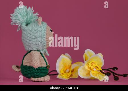 Poupée chaton Amigurumi sur fond rose à côté des fleurs d'orchidées jaunes. Un jouet de bricolage doux en coton. Un chat brun portant une combinaison verte et drôle Banque D'Images