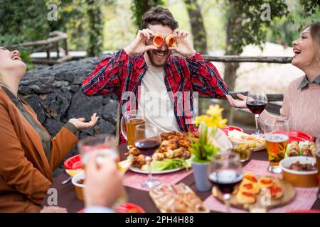Un jeune homme apporte le rire à la table en plaçant de petites tranches de pizza sur ses yeux pendant un déjeuner avec des amis. Nourriture et amitié, famil Banque D'Images