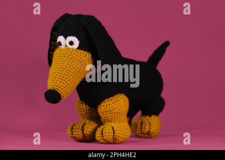 Un dachshund noir et rouge crochetté de laine naturelle et de coton, art fait à la main Poupée de chien Amigurumi sur fond rose. Un jouet de bricolage doux en coton Banque D'Images