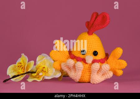 Coq orange avec peigne rouge vif sur la tête et bec crocheté, art fait à la main Poupée de poulet Amigurumi sur fond rose avec fleurs d'orchidées jaunes. S Banque D'Images