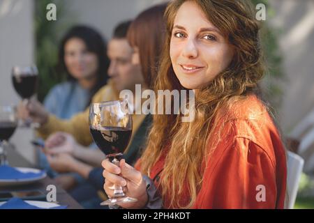 Une jeune femme caucasienne dans ses 20s-30s souriant et tenant un verre de vin - ses amis parlent et blague autour de l'arrière-plan - ils sont assis Banque D'Images