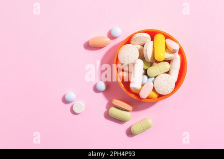 Assortiment de pilules colorées, suppléments et vitamines dans un bol orange vif disposé sur fond rose avec espace de copie. Concept de soins de santé. Banque D'Images