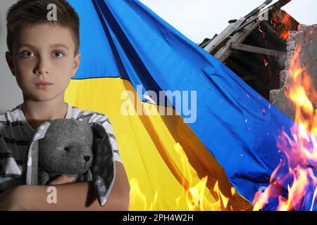 Triste petit garçon avec jouet, drapeau national et maison ruinée sur le feu. Arrêter la guerre en Ukraine Banque D'Images