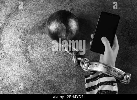 Vue de dessus de l'Internet Addicted femme dans les vêtements de prisonnier avec le ballon et la chaîne sur sa main en utilisant le smartphone à la table grise, espace pour le texte. Noir et blanc Banque D'Images