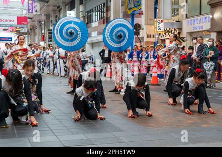 Une équipe d'enfants de danseurs de Yosakoi portant des tuniques yukata noir et blanc tout en dansant au milieu d'une rue commerçante couverte, Sunroad Shinshigai, à Kumamoto pendant le festival annuel de danse Kyusyu gassai au printemps. En arrière-plan, deux danseurs sont des parasols bleus et blancs. Banque D'Images