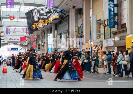 Une équipe de jeunes danseurs japonais de Yosakoi adulte portant des tuniques yukata colorées tout en dansant au milieu d'une rue commerçante couverte, Sunroad Shinshigai, à Kumamoto, pendant le festival annuel de danse Kyusyu gassai au printemps. Certains spectateurs regardent. Banque D'Images