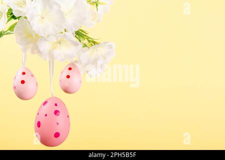 Représentation charmante de la joie de Pâques, avec trois œufs roses suspendus avec élégance d'une branche en fleur sur fond jaune pastel Banque D'Images