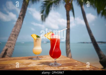 Deux cocktails sous les palmiers sur la belle plage. Singapore Sling et Pina Colada se trouvent sur une table en bois. Banque D'Images