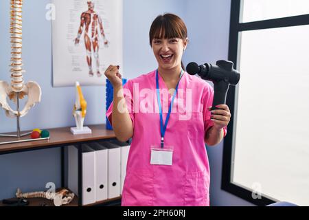 Jeune femme brune tenant un pistolet de massage thérapeutique au centre de bien-être criant fier, célébrant la victoire et le succès très excité avec le bras relevé Banque D'Images
