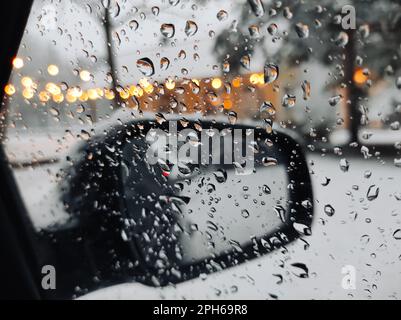 Gros plan des gouttelettes d'eau sur la vitre latérale de la voiture pendant la neige en hiver, sur fond de rétroviseur et de bokeh des feux de rue. Banque D'Images