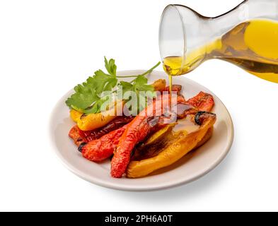 Tranches de poivrons jaunes et rouges rôtis avec feuilles de persil dans un plat blanc avec huile d'olive extra vierge versée à partir de la cruet en verre isolée sur blanc avec Banque D'Images