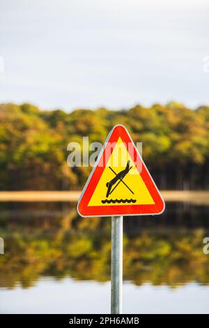 Un panneau d'avertissement jaune vif en forme de triangle interdisant la plongée dans un lac suédois paisible, soulignant l'importance de la communication Banque D'Images