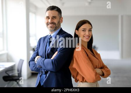 Deux hommes d'affaires à succès se tenant dos à dos avec les bras croisés et souriant à la caméra, posant dans un bureau moderne Banque D'Images