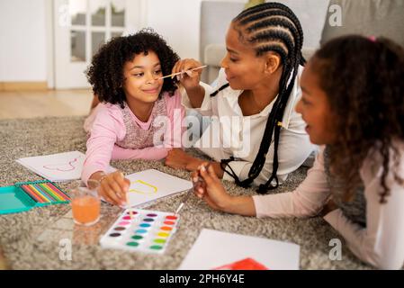 Dessin familial noir, maman faisant la peinture du visage pour les filles à l'intérieur Banque D'Images