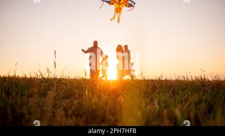 Vue panoramique sur la diversité de la famille couchant au coucher du soleil avec cerf-volant dans le ciel. Vue de dessous à travers les oreilles. Silhouette. Le père tient le jouet dans les mains Banque D'Images