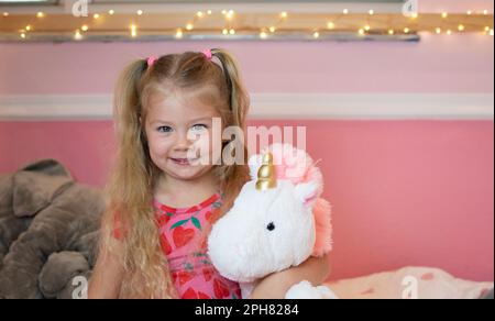 Portrait d'une petite fille blanche blonde souriante dans sa chambre rose avec un jouet en peluche licorne Banque D'Images