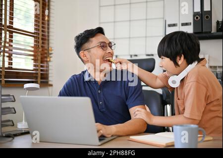 Petit garçon mignon nourrissant un cookie à son père pendant qu'il travaille dans son bureau à la maison. bon garçon, bon comportement, enfance. Banque D'Images