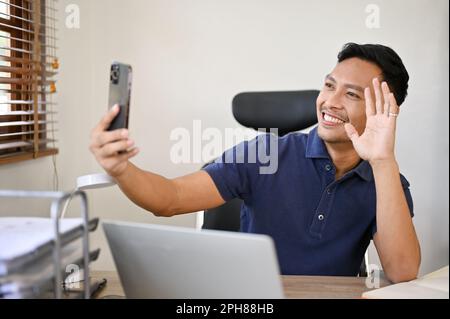 Homme d'affaires asiatique joyeux et joyeux du millénaire ayant un appel vidéo avec sa petite amie tout en étant assis à son bureau. Banque D'Images