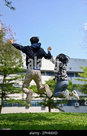 Les étudiants asiatiques insouciants et débordés de joie tiennent les mains et sautent pour célébrer leur réussite scolaire ensemble au parc du campus. retour v Banque D'Images