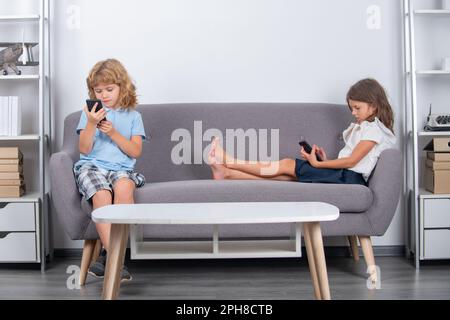 Enfants seuls avec téléphone à la maison. Des enfants heureux et joyeux qui regardent des contenus multimédias sur leur smartphone, font des appels vidéo à leurs parents, jouent à des jeux en ligne, utilisent Internet Banque D'Images