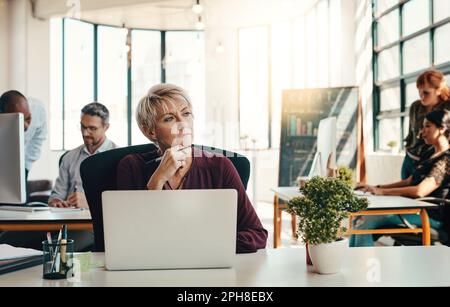 L'esprit créatif ne cesse jamais de créer. une femme d'affaires mûre et réfléchie utilisant un ordinateur portable dans un bureau moderne avec ses collègues en arrière-plan. Banque D'Images