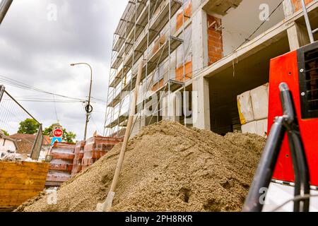 Grande colline de gravier avec pelle à outils de creusement verticale bloquée, en arrière-plan est un édifice de logement inachevé avec échafaudage, travaux en cours. Banque D'Images