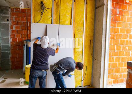 Le travail d'équipe, les réparateurs mesurent avec un mètre à ruban, en vérifiant les dimensions des murs avant d'installer les plaques de plâtre sur le mur isolé de la cloison. B Banque D'Images