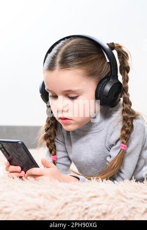 Ivano-Frankivsk, Ukraine 28 janvier 2022: Petite fille attrayante utilise des écouteurs sans fil jbl et xiomi téléphone mobile, portrait de petite fille couché o Banque D'Images