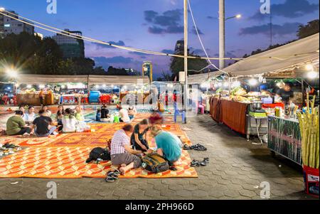 Phnom Penh,Cambodge-23 décembre 2022:comme le crépuscule tombe, les voyageurs Khmers et occidentaux s'assoient en petits groupes sur des tapis, dans une grande zone, entouré par des stands de nourriture Banque D'Images