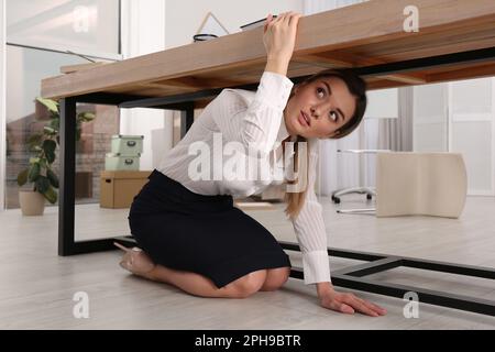 Une jeune femme effrayée se cachant sous le bureau pendant le tremblement de terre Banque D'Images