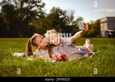 Couple de filles d'amis de l'adolescence fait selfie sur le téléphone grimacing. Les bûcherons d'école portant des chemises décontractées font des bûcherons souriants et appréciant de se reposer sur la loi du parc Banque D'Images