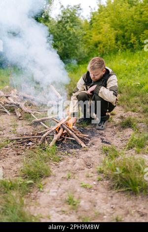 Vue de face d'un homme survivante congelé en imperméable mettant du bois de chauffage sur feu de camp à faire feu sur la rive de la rivière le soir avant le coucher du soleil. Banque D'Images