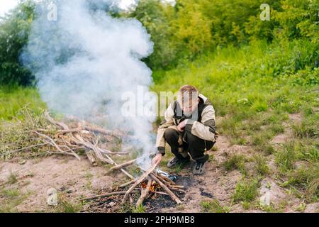 Vue de face d'un homme survivante congelé en imperméable mettant du bois de chauffage sur feu de camp à faire feu sur la rive de la rivière le soir avant le coucher du soleil. Banque D'Images