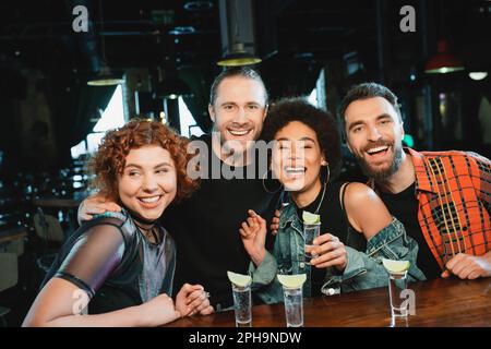 Joyeux amis interraciaux regardant l'appareil photo près des verres avec tequila dans le bar, image de stock Banque D'Images