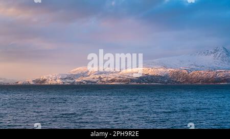 Morgenstimmung auf der Insel Senja und Kvaløya im Winter in Norwegen. DAS Morgenrot färbt schneebematte Berge und Wolken rötlich, lits Haus am Ufer Banque D'Images