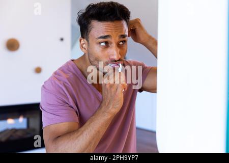 Homme biracial se regardant dans le miroir et se brossant les dents dans la salle de bains Banque D'Images