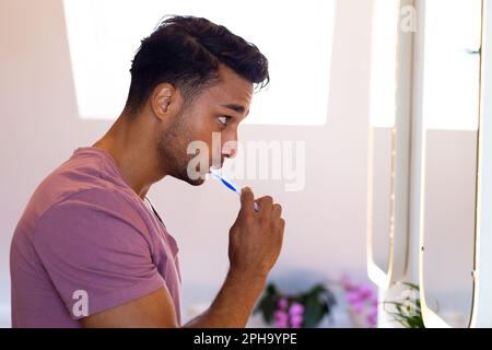 Homme biracial regardant dans le miroir et se brossant les dents dans la salle de bains Banque D'Images