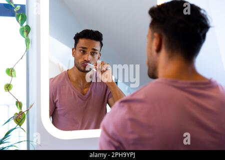 Homme biracial se regardant dans le miroir et se brossant les dents dans la salle de bains Banque D'Images