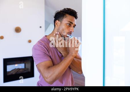 Homme biracial se regardant dans le miroir et se brossant les dents dans la salle de bains Banque D'Images