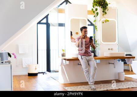 Heureux biracial homme se brossant les dents et en utilisant le smartphone dans la salle de bains Banque D'Images