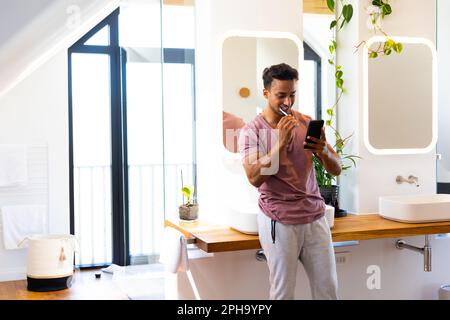 Heureux biracial homme se brossant les dents et en utilisant le smartphone dans la salle de bains Banque D'Images