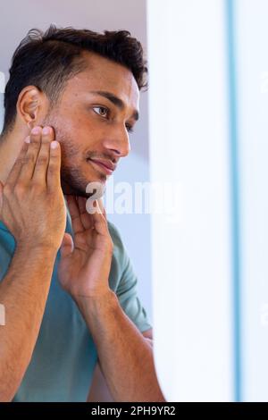 Homme biracial souriant se regardant dans le miroir de salle de bains et appliquant la crème du visage Banque D'Images